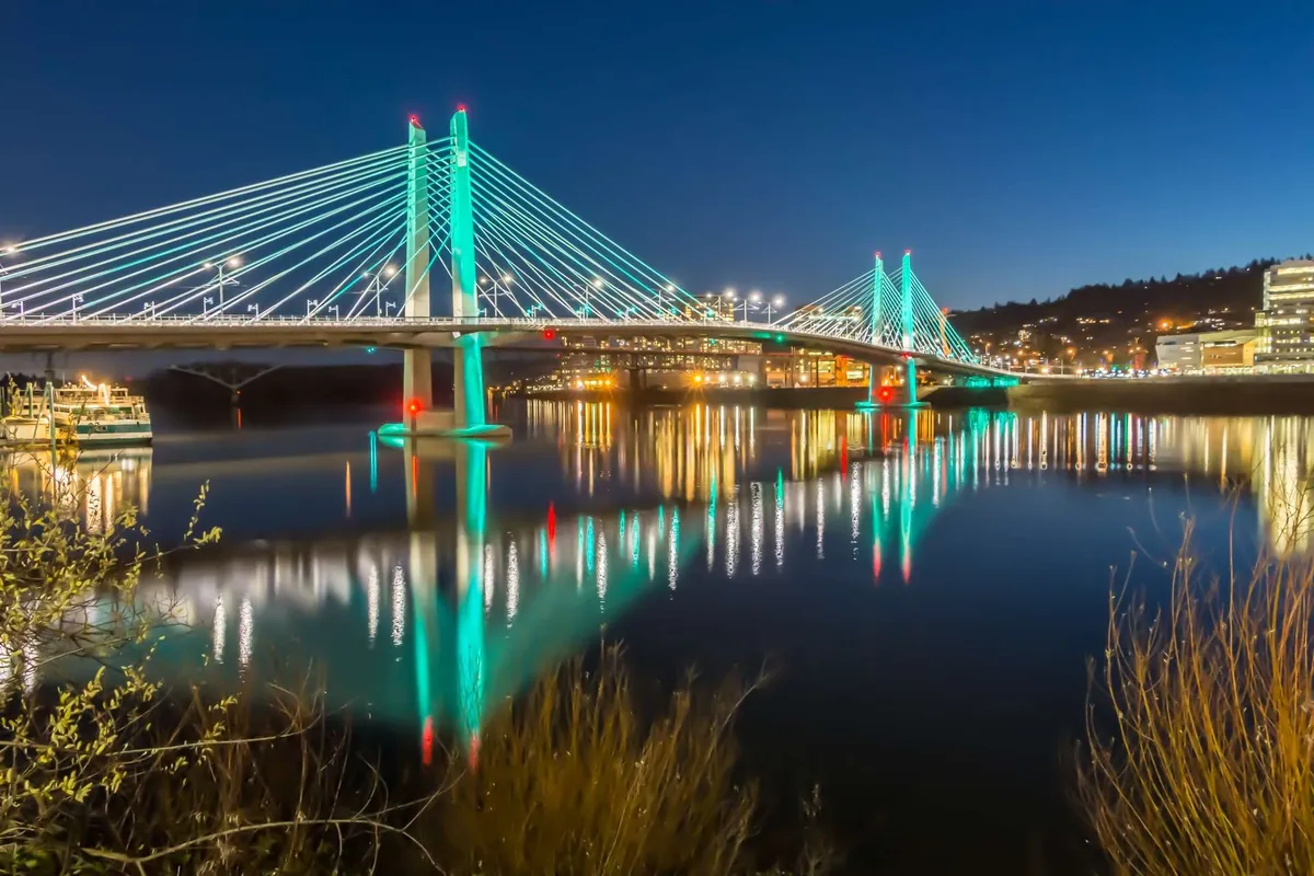 A striking nighttime view of an illuminated bridge reflecting in the water, framed by city lights. The scene captures the vibrant energy and enchanting atmosphere of the city, perfect for an evening out or taking in iconic local sights.