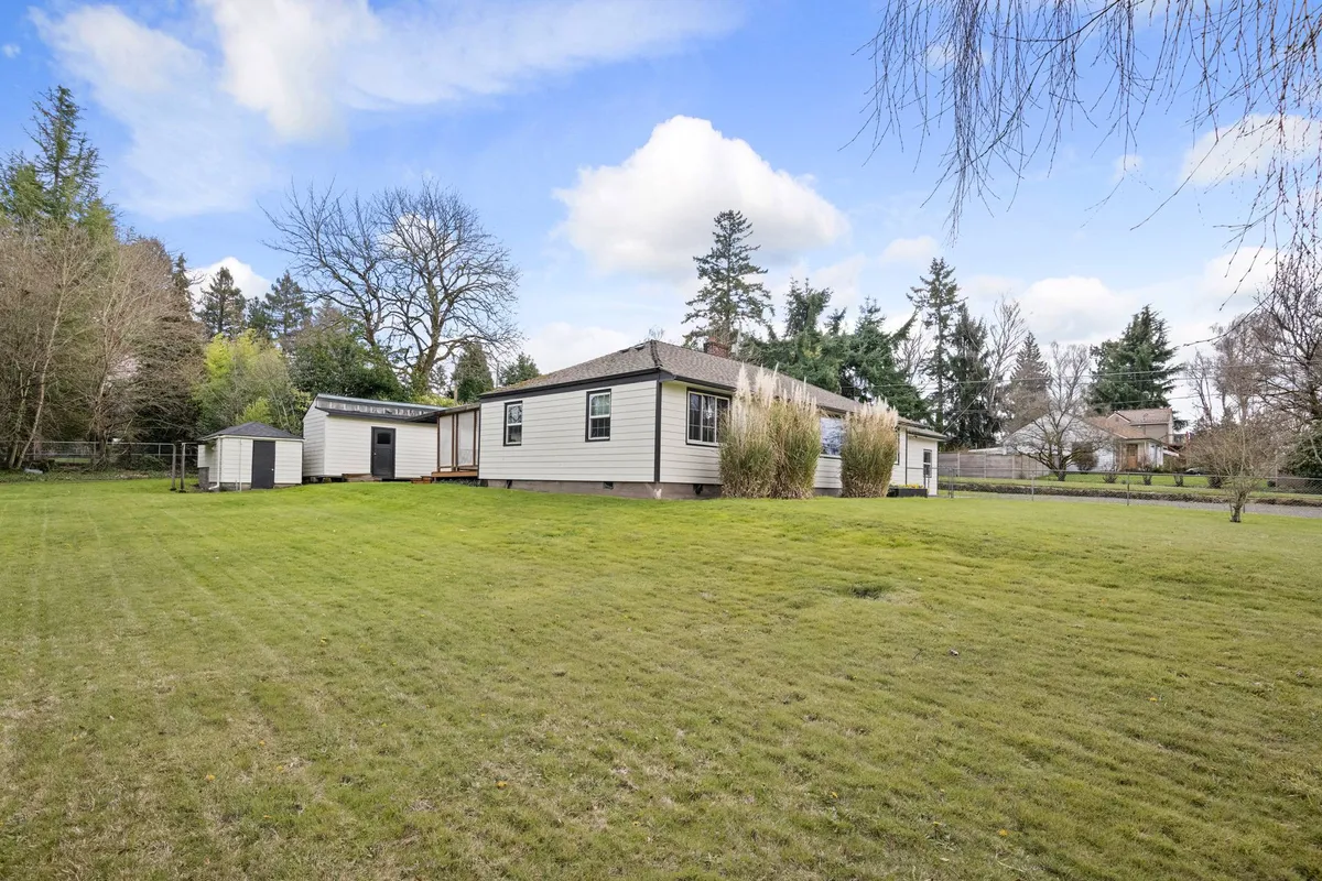 This exterior shot captures the house set amidst a generous grassy yard, fringed by mature trees and showcasing a visible garden shed. The fenced perimeter is apparent, suggesting both security and seclusion. The open space is well-suited for children or pets to play safely, while the backdrop implies ample room for outdoor activities and gatherings. The lighting evokes a peaceful, inviting mood, hinting at both sunrise mornings and sunset evenings.