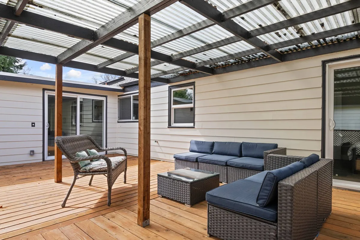 This image presents a stylish covered patio area, featuring woven outdoor furniture set atop wooden decking under a pergola. The arrangement encourages gatherings, meals, or solitary relaxation, and the structure provides shelter for all-weather enjoyment. The setting is lush and inviting, seamlessly connecting with the outdoor space beyond.