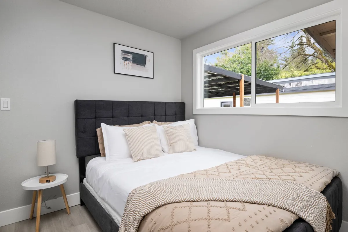 A warm, inviting bedroom with a neatly made bed decorated with plush pillows, a large window letting in ample natural light, and elegant framed artwork on the wall. The space radiates quiet, restful energy.