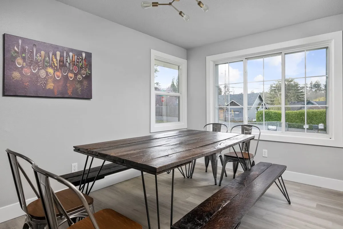 A warm dining room featuring a wooden table surrounded by metal chairs, with artwork on the wall and a large window letting in ample daylight. The inviting setting is perfect for shared meals and gatherings.