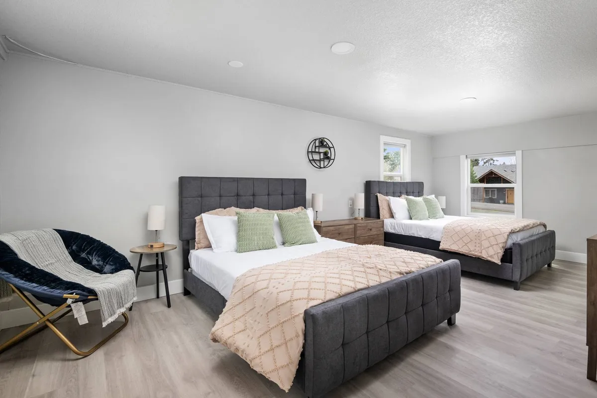 A spacious bedroom showcasing two beds, each with crisp, inviting linens. The décor is calm and understated, with a decorative wall clock and a comfortable chair by the window, creating a peaceful, shared space ideal for friends or siblings.