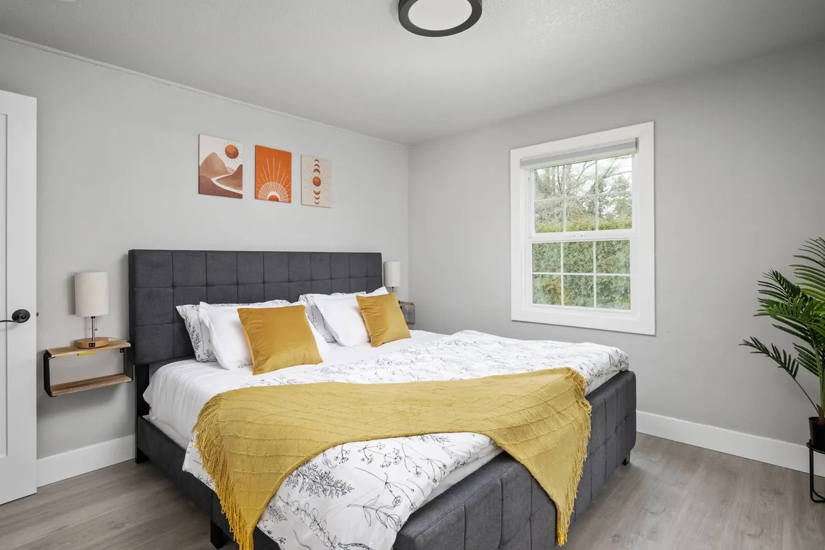 A tranquil bedroom scene featuring a comfortable bed adorned with a yellow throw blanket, artwork above the headboard, and a window framing lush greenery outside. The natural morning light fills the room, enhancing the soothing neutral palette and soft textiles.