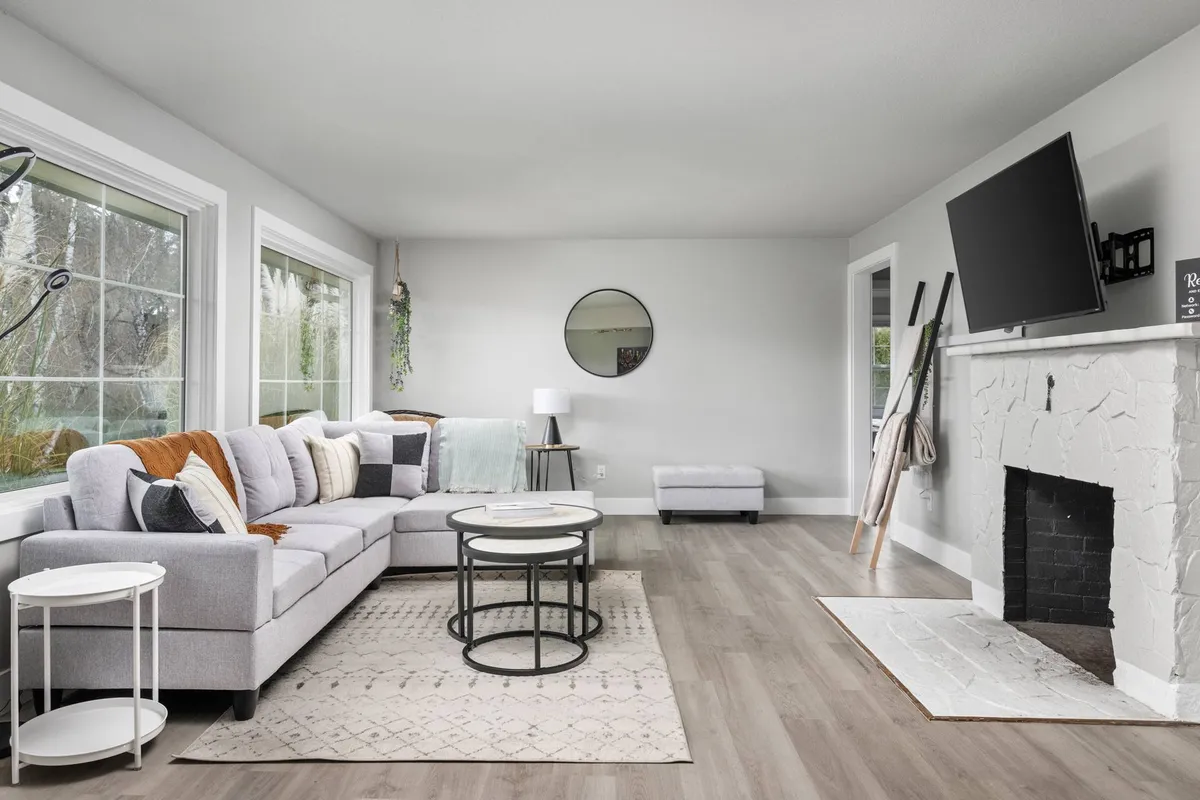 A spacious and inviting living room featuring a plush sofa, a stylish round mirror above a modern fireplace, large windows that flood the space with natural light, and a cozy coffee table—perfect for gatherings or quiet relaxation.